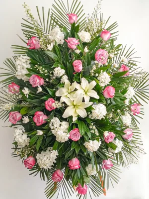 Pedestal fúnebre ovalado con rosas rosadas, hortensias y lirios blancos entre hojas de palma