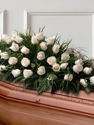 Cubre cofre alargado con rosas blancas entre follaje verde tipo hierba, dispuesto sobre un ataúd de madera