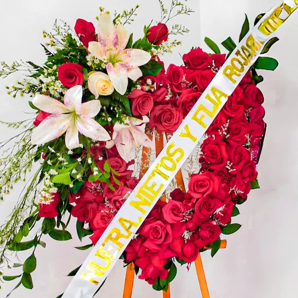 Corona Fúnebre Amor Eterno en forma de corazón de rosas rojas con un ramo de lirios blancos y rosados y cinta de condolencia, sobre atril de madera.