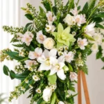 Detalle de la Corona Fúnebre Calma de Dios: lirios blancos, astromelias blancas con toques rosados, una orquídea verde, rosas crema y margaritas pequeñas entre follaje.