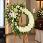 Corona Fúnebre Calma de Dios sobre atril de madera en una sala de velación: crisantemos blancos con un arreglo lateral de lirios, astromelias, rosas crema y una orquídea verde, junto al cofre y una cruz.