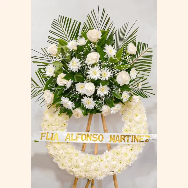 Corona Fúnebre Suspiro circular de crisantemos blancos con un copete de rosas y margaritas blancas entre hojas de palma, con cinta de condolencia.