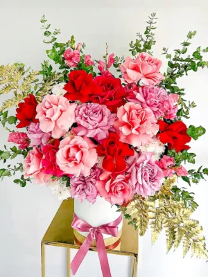 Ramo de flores con rosas rojas y rosadas, follaje verde y dorado en base blanca elegante, ideal para expresar amor y admiración.