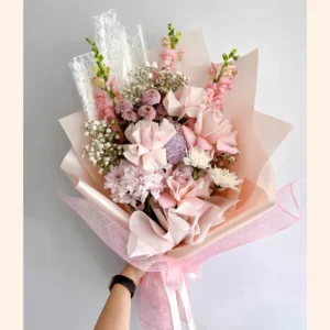 Bouquet envuelto en papel rosa con rosas rosadas y blancas, boca de dragón rosa y flor de nube, lazo de malla rosa.