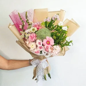 Bouquet de Flores Fleurir con gerberas rosadas, rosas, margaritas y un pompón verde, en papel beige con lazo de encaje.