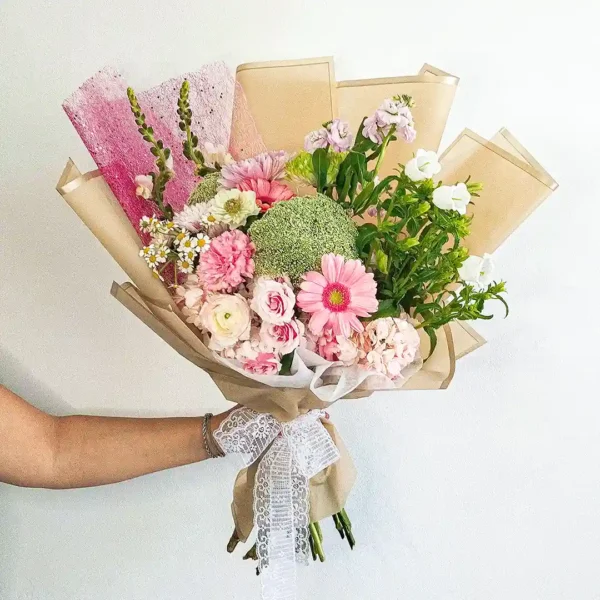 Bouquet de Flores Fleurir con gerberas rosadas, rosas, margaritas y un pompón verde, en papel beige con lazo de encaje.