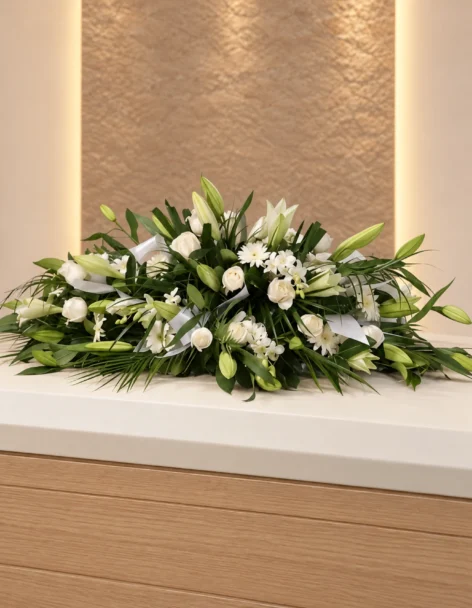 Cubre cofre de rosas blancas, lirios, gerberas blancas y orquídeas con follaje verde y cintas blancas, sobre un cofre de madera.