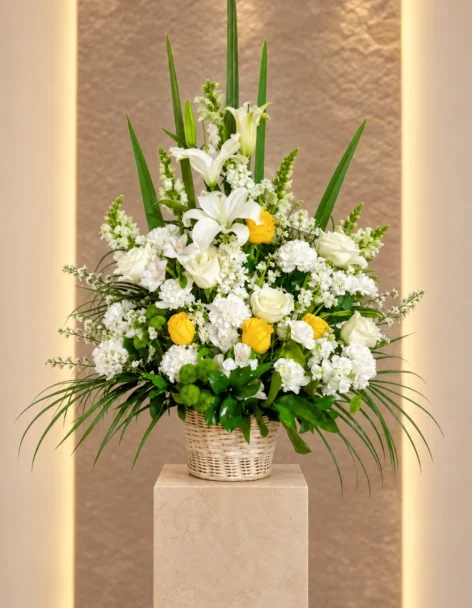 Ramo fúnebre en canasta de mimbre con lirios y rosas blancas, rosas amarillas, claveles blancos, boca de dragón y pompones verdes, sobre un pedestal de mármol.